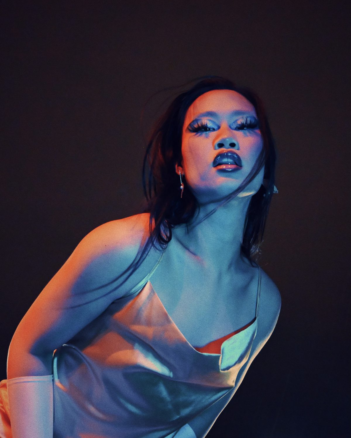 Performer Kylie Mooncakes poses for the camera. Long black hair falling on their shoulders, long eye lashes, lush red lips and wearing a gold dress, Kylie is lit by contrasting red and blue lights in front of a black background.