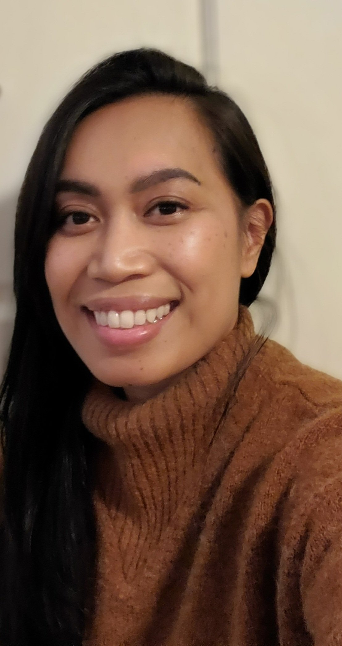 Lea wears a chestnut sweater against a white backdrop. She has long black hair, brown skin, and a wide smile, showing her teeth. 
