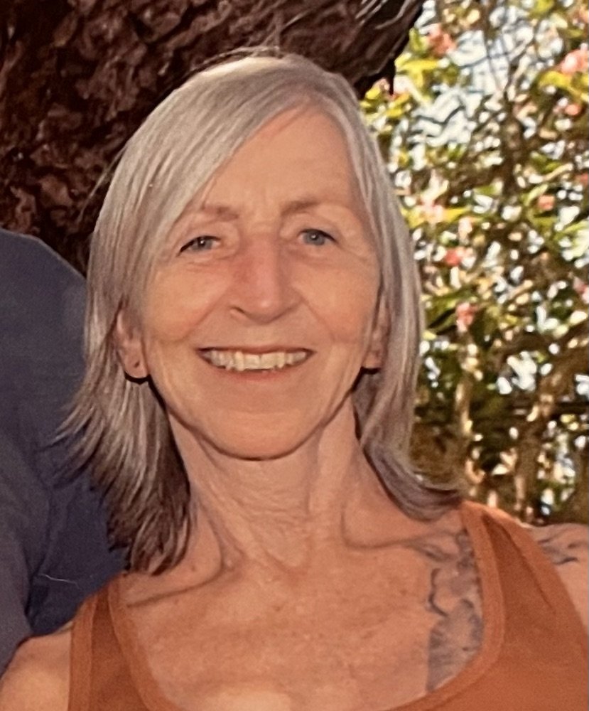 Tracee is smiling with a leafy tree in the background. She has shoulder length grey hair and is wearing a tank top.