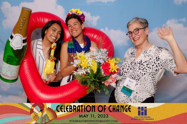 Standing in a photobooth with blue sky background, Merril waves, holding a large beautiful bouquet. In a heart-shaped inflatable, Amarinthia and Lea hold a fake inflated champagne bottle, a rubber chicken, and wear a fake flower head dress.
